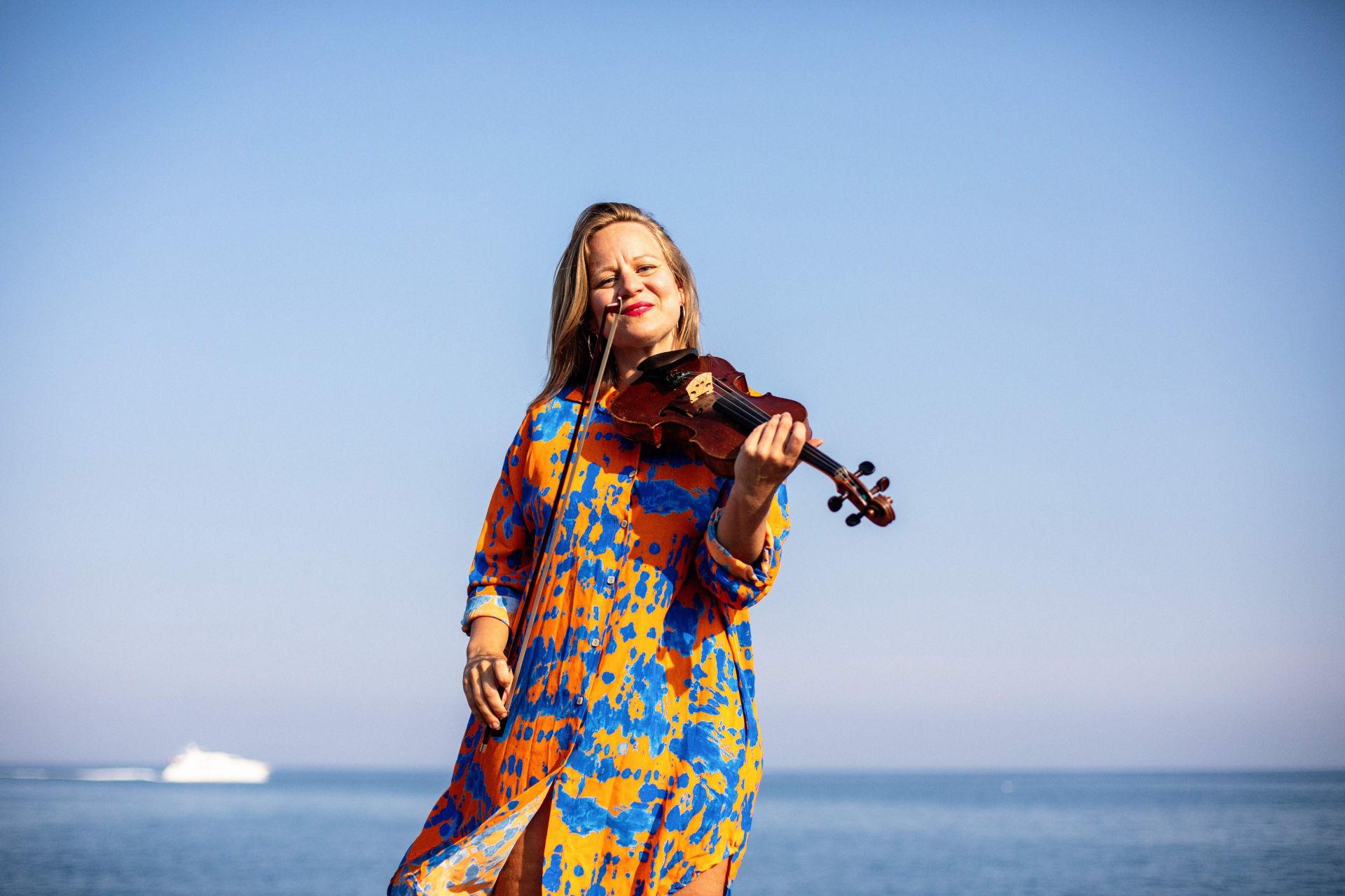 Pedagog och konstnär Lina Teir står vid en strand i en färgglad klänning. Lina håller i sin famn en fiol. Lina tittar rakt in i kameran och ler.