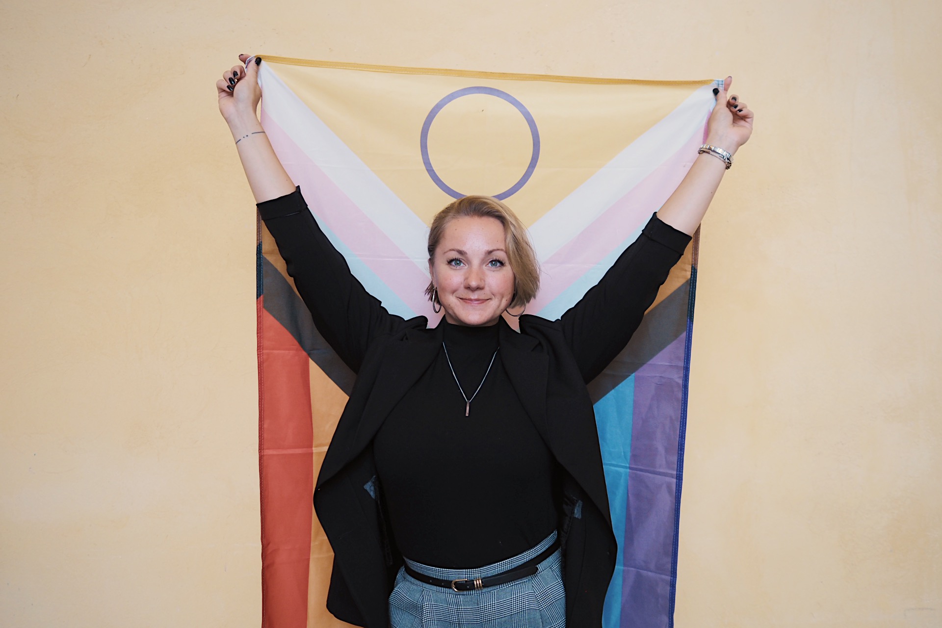 Fotografi i färg av Isabel Flemming, verksamhetsledare för Regnbågsalliansen. Isabel håller uppe en Progress Pride-flagga med båda händerna.