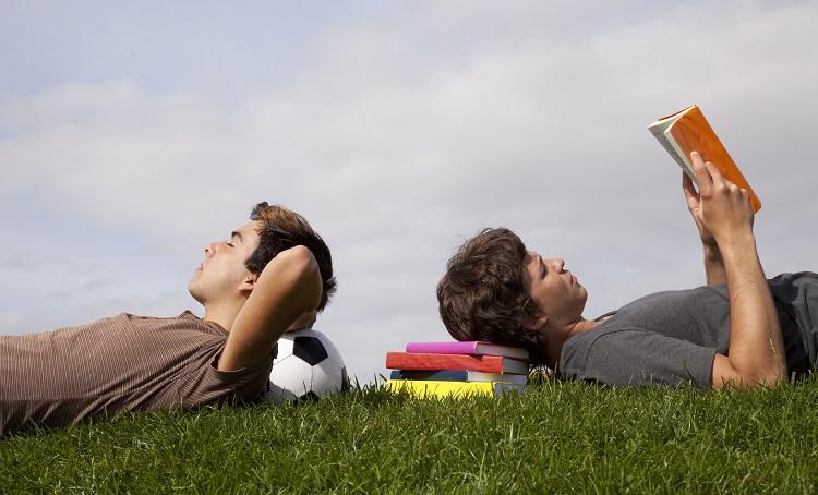 Två män ligger på en gräsmatta, huvudena mot varandra. Den ena med huvudet på en fotboll, den andra med huvudet på en trave böcker, läsande en bok.