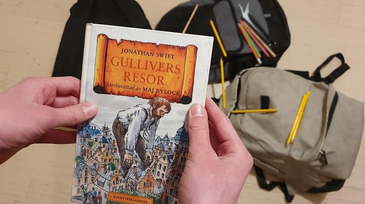 Händer som håller i boken Gullivers resor. I bakgrunden ligger två ryggsäckar.