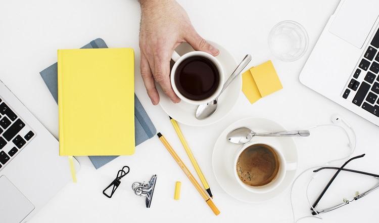 stämningsskapande bild med gula detaljer, pennor, papper, glasögon, en hand som håller i en kaffekopp.