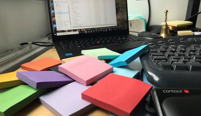 Ett skrivbord med dator, tangentbord och mängder av post it-lappar i olika färger.