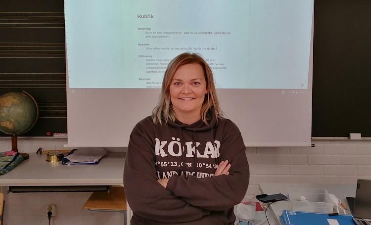 Lärare Cecilia Österlund i klassrummet. I bakgrunden ser man en presentation av en labbrapport.